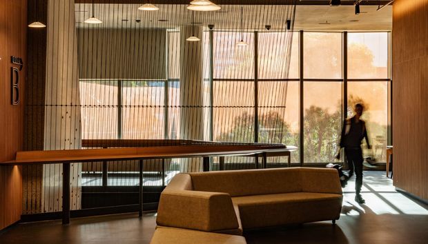 A warm, contemporary reception or lounge space with extensive floor-to-ceiling glazing, vertical timber detailing, pendant lighting, a long timber counter or table, and a substantial upholstered sofa. The interior has a refined, softly lit atmosphere with views of greenery beyond the glass.