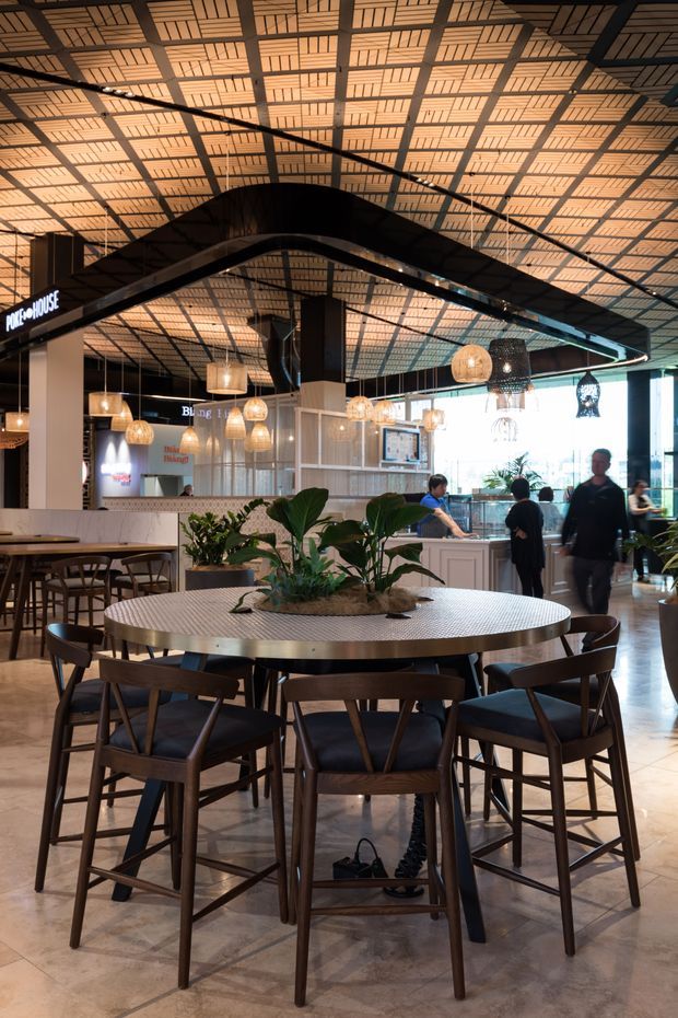 A spacious contemporary commercial dining area or food hall with a large round communal table surrounded by dark timber chairs. The interior features a patterned warm-toned ceiling with exposed dark structural framing, pendant lights in woven and glass forms, polished concrete or stone-look flooring, white service counters, indoor plants, and visible food or beverage outlets in the background. The atmosphere is warm, inviting, and high-volume, with people moving through the space.