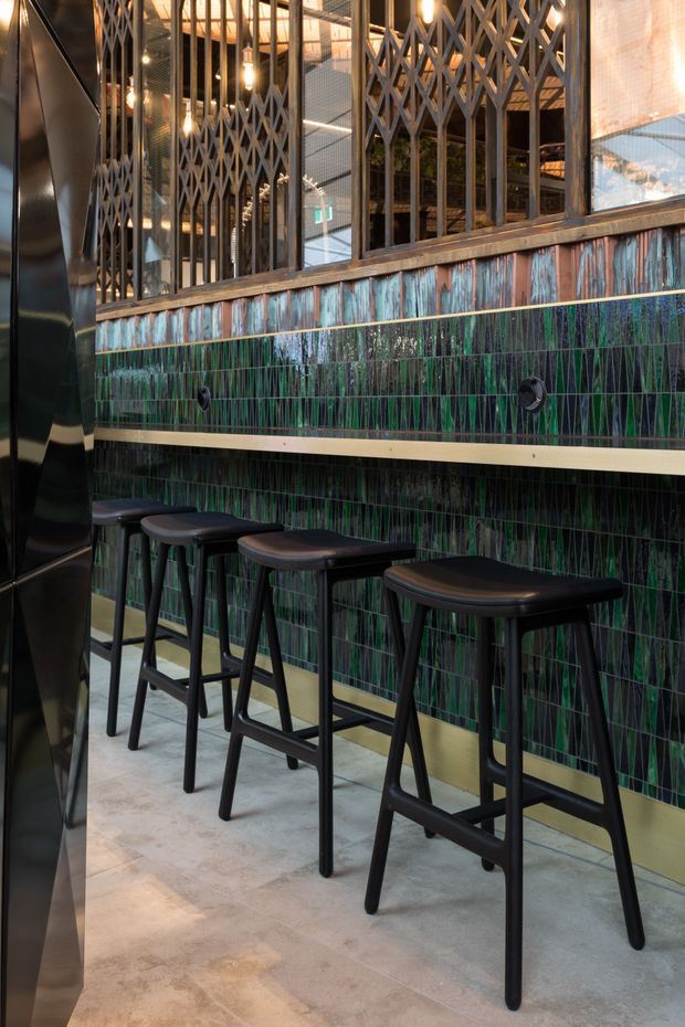 A moody commercial hospitality interior showing a narrow high-seating counter lined with black bar stools. The counter wall is clad in vertically patterned dark green and blue tiles with pale grout and brass-toned horizontal trims. Above it, black metal-framed glazing and diamond-pattern security screens reveal an industrial-style interior with exposed structure and warm pendant lighting. The floor appears to be pale stone or concrete-look tile, while a glossy dark partition frames the left side.
