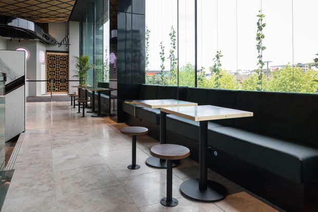 A contemporary commercial dining or café interior with a long upholstered banquette beside floor-to-ceiling windows. Small square tables with pedestal bases and stools line the seating area, while a patterned ceiling, polished stone-look flooring, dark wall panels, and a decorative geometric timber screen add visual interest. Bright greenery and an urban setting are visible through the glazing.