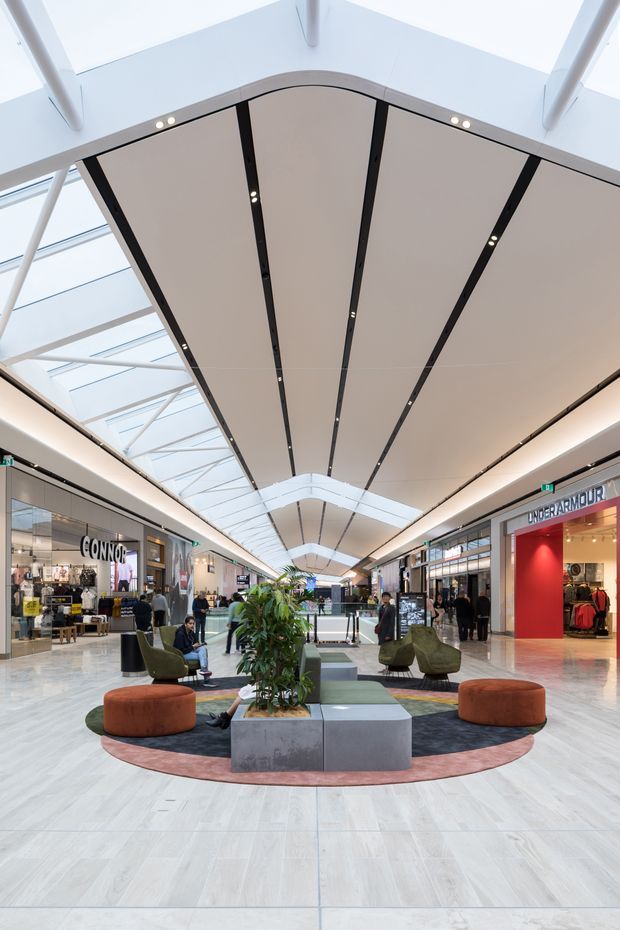 Interior view of a bright contemporary shopping mall atrium, looking along a retail concourse with storefronts on both sides. The space features a large segmented suspended ceiling with integrated linear downlights, extensive skylighting and exposed white structural framing. A central circular seating and planting zone includes upholstered lounge chairs, rounded ottomans, patterned rugs, and concrete planters. Retail signage and shoppers are visible in the background.