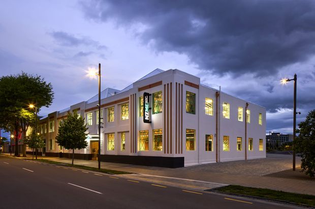 Twilight street view of a prominent two-storey contemporary commercial building with a white rendered facade, strong vertical geometric detailing, illuminated multi-pane windows, and a dark base. The building fronts a paved sidewalk and roadway, with dramatic cloudy skies and street lighting.