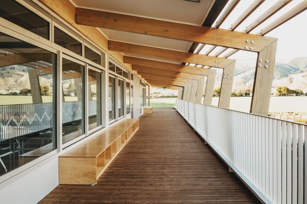 A long, covered exterior walkway or balcony with warm timber decking, exposed timber beams and rafters, extensive glazing and built-in timber bench seating along one side. A white balustrade runs along the open edge, with a bright mountainous landscape visible beyond. The linear structure creates a strong covered outdoor circulation space with repeating timber frames.