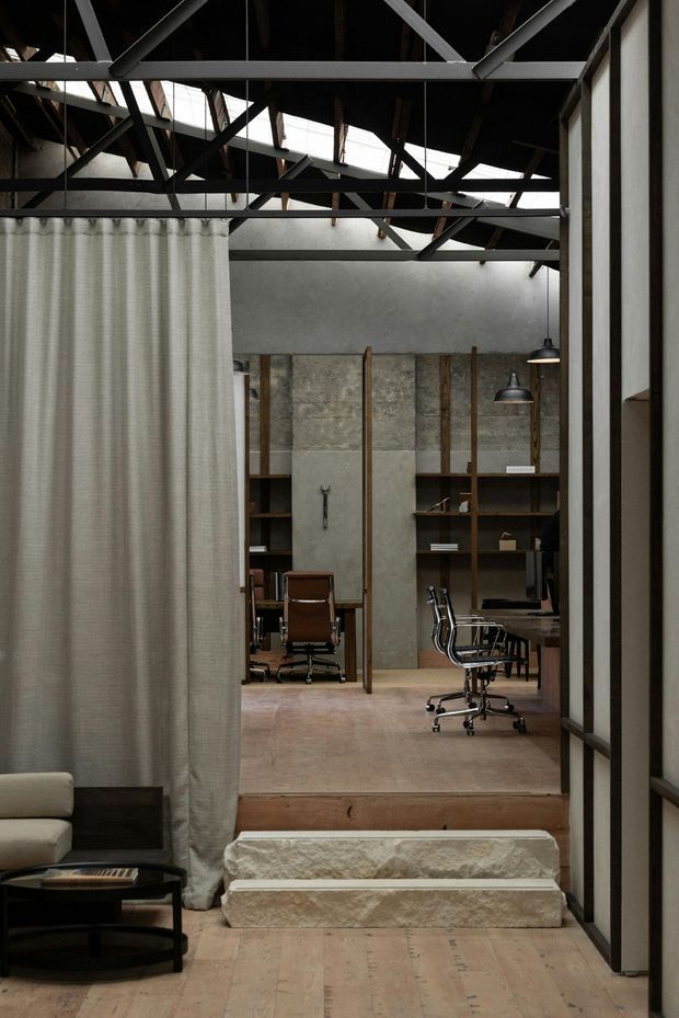 Interior view of a converted industrial-style workspace opening beyond a small raised seating area. The space features exposed dark steel roof trusses and rafters, skylights, raw textured concrete or stone wall panels, timber shelving and partitions, polished concrete or stone steps, and muted neutral finishes. A curtain partially screens a lounge area in the foreground, while office desks and wheeled chairs are visible in the workspace beyond.