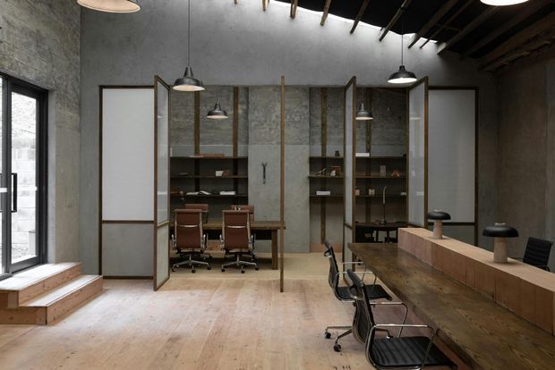 A spacious industrial-style commercial interior resembling a creative studio or office. The room features exposed concrete walls, a timber floor and ceiling structure, black pendant lights, timber-framed glazed partitions, open shelving, workstations, and a long timber communal counter with chairs. Natural light enters through tall windows on the left.