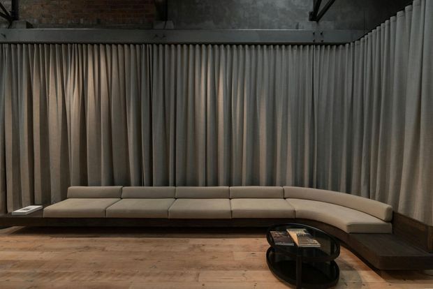 A dark, industrial-style lounge featuring a long curved upholstered sectional sofa set against full-height pleated grey curtains. The room has exposed brick and concrete surfaces, black structural framing, dark timber skirting, a warm timber floor, and a low round black glass coffee table with books.