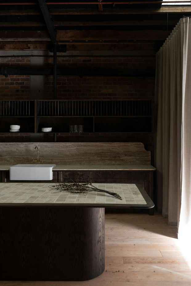 A moody, dark-toned kitchen interior in an industrial adaptive-reuse style. The space features exposed red brick walls, dark timber ceiling beams and cabinetry, open shelving, a large island with a pale greenish tiled surface, a white rectangular farmhouse-style sink with brass tapware, and a long stone or travertine-look benchtop and splashback. Floor-to-ceiling beige curtains soften the room beside a bright window, while timber flooring adds warmth.
