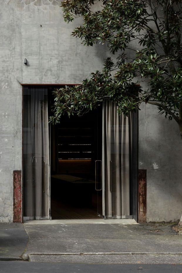 A restrained urban building entrance set into a tall raw grey concrete facade. Full-height glazed double doors with dark metal frames and long vertical handles are partly covered by sheer curtains, revealing a dim interior beyond. Weathered reddish masonry fragments flank the doorway, while a mature leafy tree partially obscures the upper right portion of the facade. The scene has a moody, minimalist industrial character.