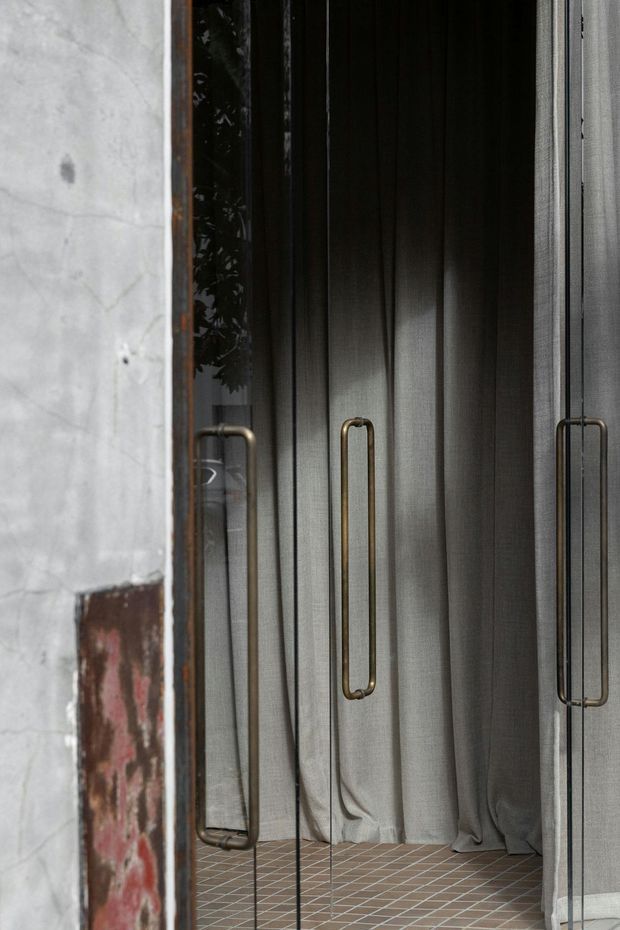 Close-up view of a refined dressing or wardrobe area framed by a raw concrete and exposed brick wall. Dark bronze-framed glazed doors with elongated brass-toned pull handles open onto a space lined with charcoal-grey curtains and finished with small brown rectangular floor tiles. Reflections in the glazing reveal the surrounding doorway and foliage. The composition contrasts industrial masonry with tailored, luxurious wardrobe detailing.