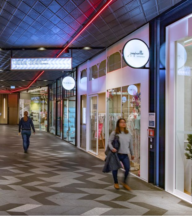 Interior view of a contemporary retail shopping arcade or commercial mall corridor. The space features a patterned geometric tiled floor, dark decorative pressed ceiling panels, illuminated red linear lighting, glazed shopfronts, retail signage, and a mix of black, white, and pastel storefront finishes. Several people are walking through the corridor.