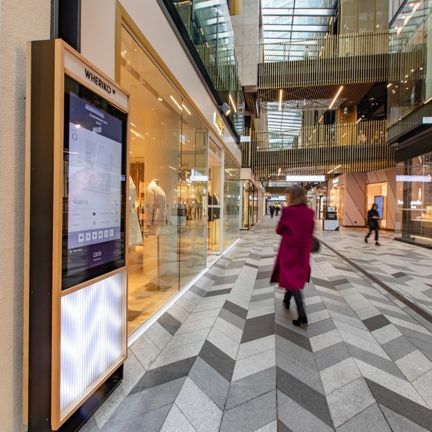 A bright, contemporary indoor shopping centre atrium with retail storefronts on both sides, a patterned geometric tiled floor, illuminated digital directory signage, suspended timber-toned slatted screens, and a glazed roof bringing in natural light. Several shoppers are present in the broad circulation space.