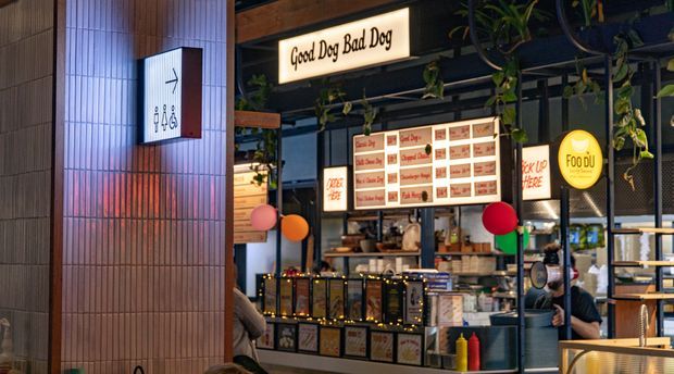 Interior view of a contemporary food-hall or casual dining venue, showing a branded counter service stall with illuminated menu boards, hanging plants, pendant-style lighting, and adjacent seating. A ribbed tiled column with directional signage occupies the foreground, while warm decorative lights and colourful balloons add visual accents.