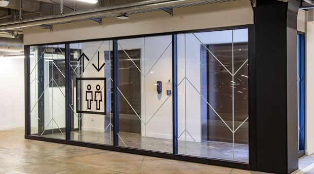 Commercial interior corridor featuring a glazed lift lobby or elevator enclosure with black-framed glass panels, geometric line decals, and an accessible restroom/elevator directional sign. The exposed services and polished concrete floor create an industrial office fit-out.