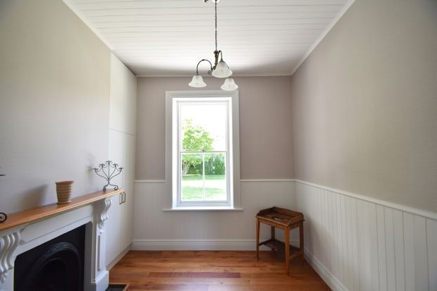 A narrow, light-filled traditional interior with warm timber flooring, pale taupe walls, white wainscoting, and a white-painted decorative fireplace surround at left. A tall sash-style window provides a garden view, while a small timber side table and vintage-style multi-light pendant reinforce the heritage character.