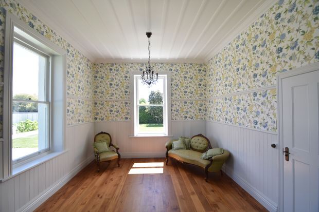 A bright, narrow traditional sitting room with floral wallpaper above white vertical wall panelling, a white beadboard-style ceiling, and warm polished timber flooring. Two upholstered vintage armchairs and a small settee sit around the room, with a delicate central chandelier. White-trimmed windows and a paneled door reinforce the period character.