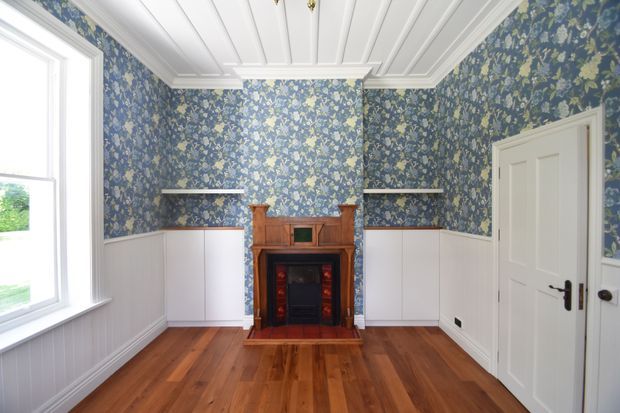 A compact, bright room with rich blue botanical wallpaper above white vertical wall panelling, a polished timber floor, and a traditional timber fireplace with a dark inset. White-painted panelled ceiling, detailed cornices, a large window, built-in white cabinetry, and a paneled door complete the refined heritage-inspired interior.