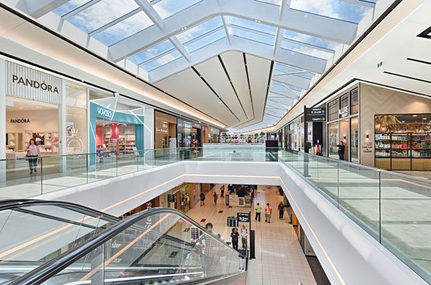 Bright, contemporary multi-level shopping mall interior viewed along an upper-floor retail concourse. The space features numerous storefronts, glass balustrades, escalators descending to a lower level, polished light flooring, and a large glazed skylight roof that fills the corridor with natural light.