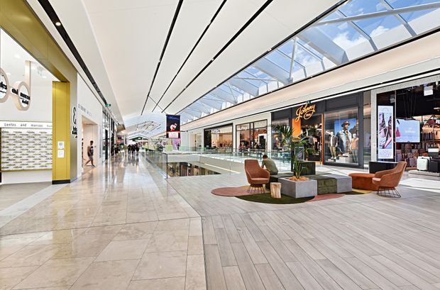 Wide-angle view of a bright contemporary shopping mall interior with a long retail corridor, polished stone-tile flooring, timber-look tiled flooring in a seating area, glass shopfronts, indoor planting, and lounge chairs around a low table. A large glazed skylight and linear ceiling panels bring abundant daylight into the atrium-like circulation space, with a visible lower-level opening and escalator area in the distance.