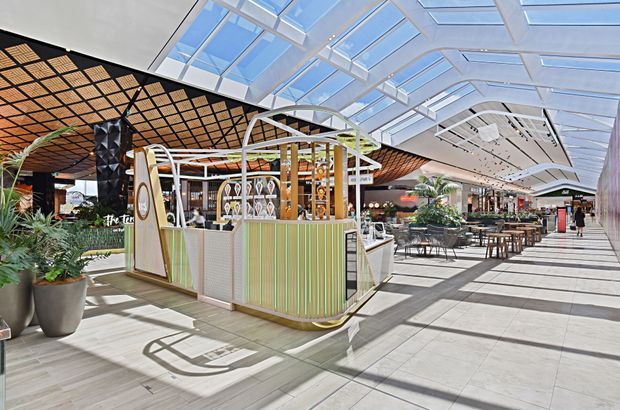 Bright contemporary indoor commercial dining and retail environment beneath a large glazed skylight. The image is dominated by a small freestanding food or beverage kiosk with rounded corners, pale green vertically ribbed panels, timber framing and decorative display shelving. Surrounding features include tiled flooring, communal dining tables and chairs, abundant indoor planting, a dramatic black-and-timber patterned ceiling, and extensive clerestory-style glazing.