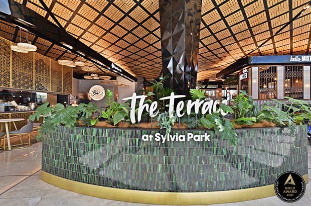 Wide interior view of a contemporary food-hall or hospitality venue at Sylvia Park, centred on a large curved planter or counter clad in vertically arranged green-toned tiles with a gold-coloured base. Dense tropical planting, patterned screens, decorative pendant lights, dark geometric structural elements, and a warm gridded timber ceiling create a lush, vibrant commercial atmosphere. Restaurant and retail-style spaces are visible in the background, with overlaid promotional text reading “The Terrace at Sylvia Park” and a gold award badge.