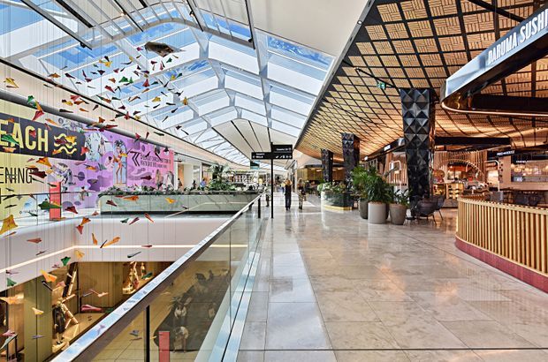 A bright, contemporary multi-level commercial interior resembling a shopping or dining precinct. The space features a broad polished stone walkway, glass balustrades overlooking a lower level, extensive skylights and glazed roof panels, colourful suspended origami-like installations, large graphic wall artwork, indoor planting, and a restaurant frontage with a warm geometric timber ceiling and decorative dark structural columns.
