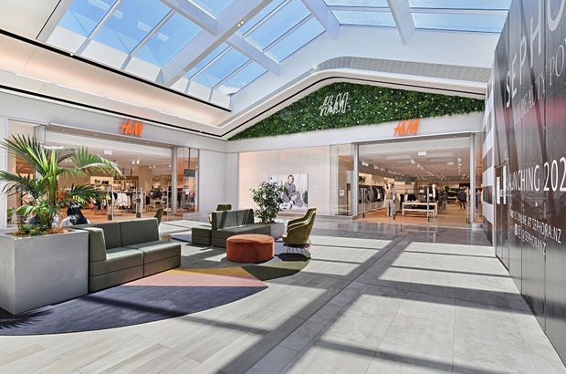 Bright contemporary shopping-centre interior with a glazed atrium roof, polished light-grey tiled flooring, retail storefronts including H&M, indoor tropical planting, and a central lounge seating area. Strong daylight and geometric skylight shadows define the spacious commercial environment.