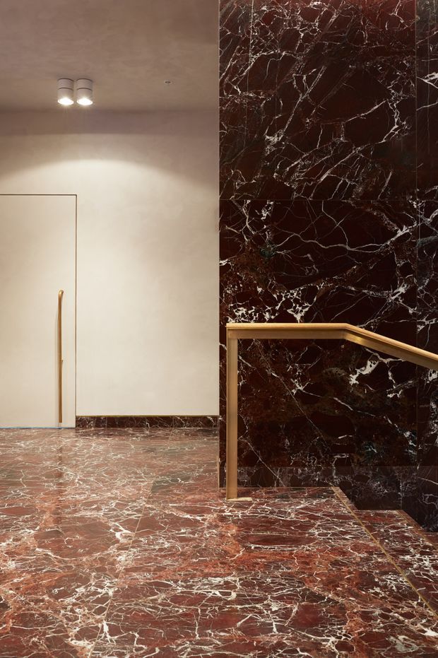 A refined interior stair landing or hallway featuring dramatic red-brown veined marble flooring and matching full-height marble wall cladding. A minimalist flush door with a slim brass handle is set into a pale plaster wall, while a sculptural brass handrail follows the stair edge. Recessed ceiling spotlights provide warm illumination, creating a luxurious, high-contrast material-focused composition.