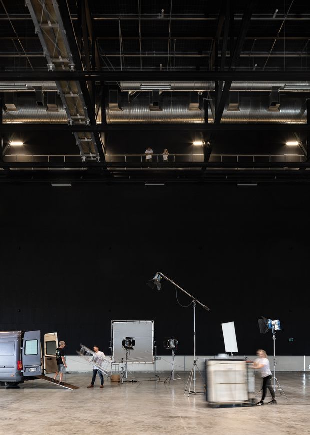 A large dark industrial-style studio or event hall with a high exposed-services ceiling, visible steel structure, suspended ductwork, overhead lighting, and a mezzanine walkway. The polished concrete floor is occupied by photographic or film-production equipment, lighting stands, a van, rolling cases, and several people. The space has a dramatic black interior and expansive open floor area.