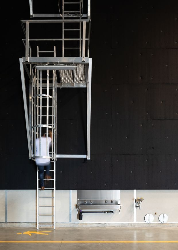 A dramatic industrial interior featuring a tall suspended metal access structure and ladders against a dark black wall. A person is climbing down the ladder, with stainless-steel wall-mounted fixtures, exposed services, and yellow floor safety markings below. The space has a utilitarian, warehouse-like character with strong vertical scale and high contrast.