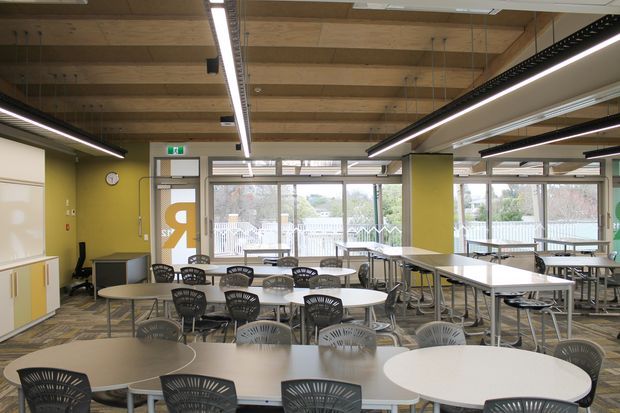 A bright, contemporary cafeteria or dining hall with numerous round and rectangular tables, black perforated chairs, and extensive glazing. The space features a warm exposed timber-beam ceiling, suspended linear LED lighting, lime-green accent walls, grey framed windows and doors, and resilient patterned flooring. Outdoor decking and planting are visible through the windows.