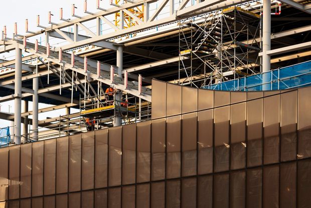 Photograph of a large commercial building under construction, showing exposed steel structural framing, extensive scaffolding, temporary edge protection, construction platforms, and brown translucent site hoarding in the foreground. Workers in high-visibility clothing are visible within the construction zone.