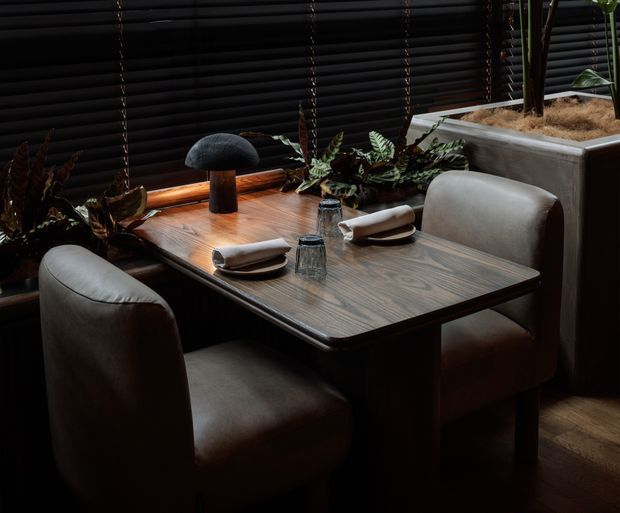 A moody, intimate dining nook with a rectangular dark timber dining table, two upholstered rounded-back chairs, sculptural mushroom-shaped table lamp, folded napkins, tableware, and lush indoor plants. Dark horizontal blinds and a raised planter with a concrete-like finish form the backdrop, while warm timber flooring and low lighting create a refined restaurant-like atmosphere.