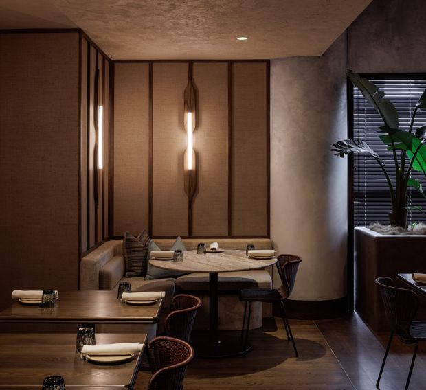 A dimly lit, refined restaurant dining area with warm timber flooring, dark textured walls, woven upholstered banquette seating, round and rectangular dining tables, and sculptural pendant wall lighting. Vertical timber-framed textured panels form a feature wall, while a large potted plant sits beside a window with dark horizontal blinds.