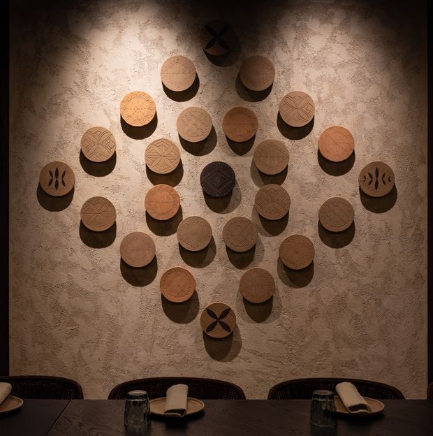 Warm, moody restaurant or hospitality interior featuring a large decorative installation of round carved wooden discs arranged on a textured plaster wall. A dark dining table with place settings and woven-backed chairs occupies the foreground, with dramatic directional lighting creating strong shadows.
