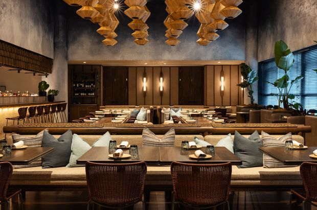 A sophisticated restaurant or hospitality dining interior with long communal dining tables, upholstered banquette seating, woven dining chairs, layered neutral textiles, and dark timber cabinetry. The space features a dramatic dark ceiling, textured concrete-look walls, warm timber panelling, tall windows with blinds, indoor plants, and sculptural clusters of suspended timber pendant lights. The atmosphere is warm, luxurious, refined, and intimate.