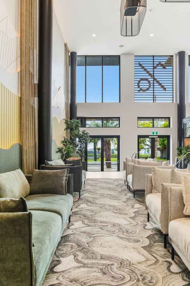 A bright double-height interior lounge or reception space with floor-to-ceiling glazing, tall black cylindrical columns, sculptural pendant lighting, and rows of upholstered lounge chairs arranged along a wide patterned carpet. A large decorative vertical feature wall with timber slats and curved forms occupies the left side, while landscaped outdoor areas are visible through the glass doors and windows.