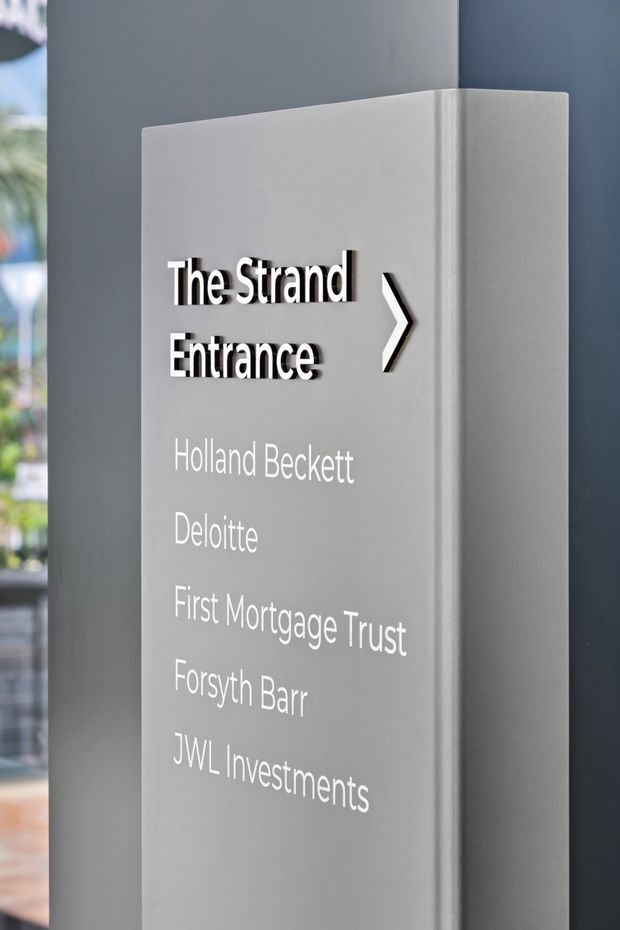 A close-up photograph of a freestanding grey wayfinding and directory sign in a contemporary commercial building lobby. The sign features raised black-and-white lettering reading “The Strand Entrance” with a directional arrow, followed by a list of office tenants. A glazed area and blurred greenery are visible at the left edge, while dark grey architectural wall surfaces frame the sign.