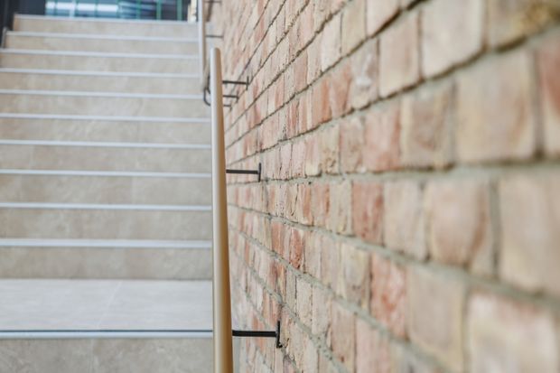 Close-up architectural view of a stairway with pale stone or concrete treads, slim metal-edged risers, a simple timber handrail, and an exposed red brick feature wall running alongside the stairs. The composition emphasizes the tactile brickwork and contemporary stair detailing.