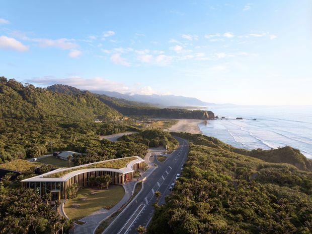 Aerial view of a low, curving contemporary building set within dense coastal vegetation beside a winding road. The image shows a broad sandy beach, rocky headland, ocean shoreline, and distant forested hills. The building has extensive glazing, a sweeping roofline, and landscaped grounds.