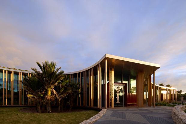 A low-rise contemporary building photographed at dusk, featuring a sweeping curved glazed facade with slender vertical structural fins and a prominent flat-roofed entrance canopy. Warm interior lighting contrasts with dark timber or metal framing, while palms and landscaped planting soften the approach. A paved pedestrian entry path leads through a low stone-edged landscape.