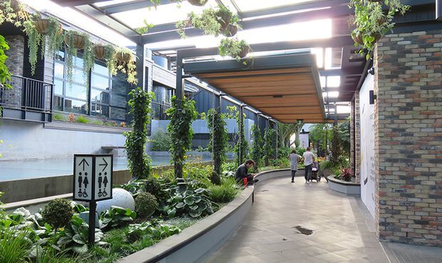 A covered landscaped pedestrian courtyard or walkway in a contemporary building complex, with extensive hanging and climbing greenery, planted garden beds, concrete paving, dark metal and glass structures, a timber-lined canopy, and exposed multicoloured brick walls. Directional restroom signage and people are visible in the distance.