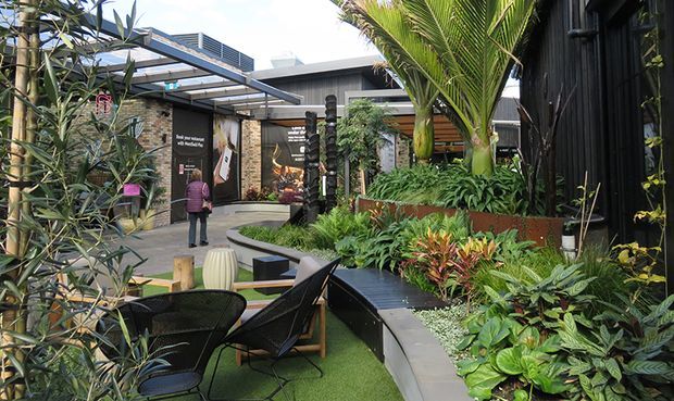 A landscaped outdoor courtyard or garden terrace with lush tropical planting, raised garden beds, dark timber-clad structures, paved walkways, outdoor seating, and a pergola-like canopy. The space has a contemporary commercial or hospitality feel, with layered greenery and integrated curved seating edges.