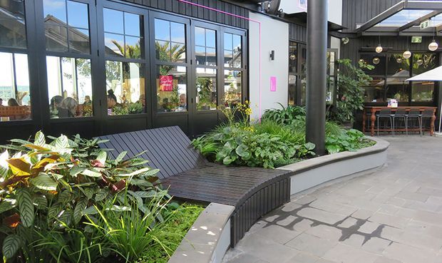 A contemporary indoor-outdoor commercial courtyard or atrium with dark metal-framed glazing, exposed structural columns and roof elements, lush integrated planting, built-in curved timber bench seating, and a paved circulation area. A café or hospitality seating area is visible in the background through the open space.