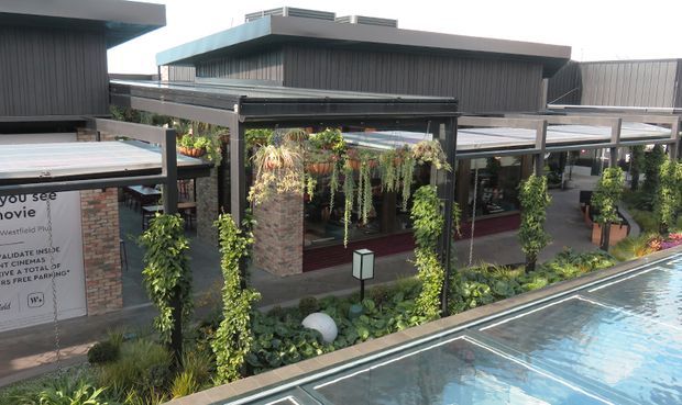 Exterior view of a contemporary commercial venue beside a swimming pool, featuring dark vertical cladding, flat roof forms, covered outdoor dining or walkway structures, hanging greenery, planted landscaping, and brick masonry walls. The pool occupies the foreground.