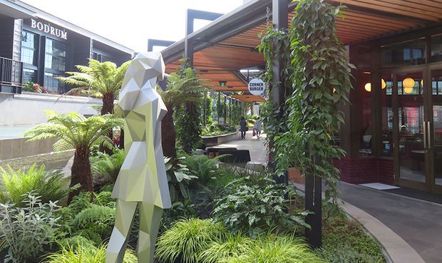 A landscaped pedestrian courtyard or outdoor dining precinct with lush tropical planting, a faceted geometric human sculpture, timber-lined covered structures, and glazed storefronts. The image includes a visible restaurant or commercial venue entrance and signage, with modern angular architecture and shaded walkways.