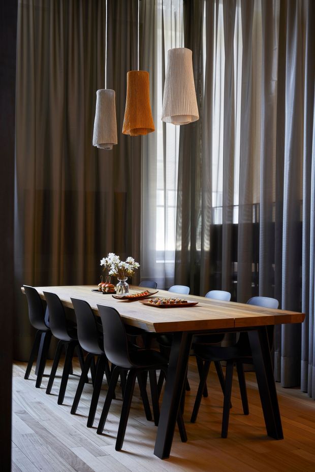A refined dining room with a long light timber dining table, black sculptural dining chairs, and three vertically ribbed pendant lights in white, amber, and beige. Floor-to-ceiling sheer and dark curtains frame a tall window, creating a dramatic, warm atmosphere. The room features timber flooring, restrained contemporary furnishings, and a small floral table arrangement.