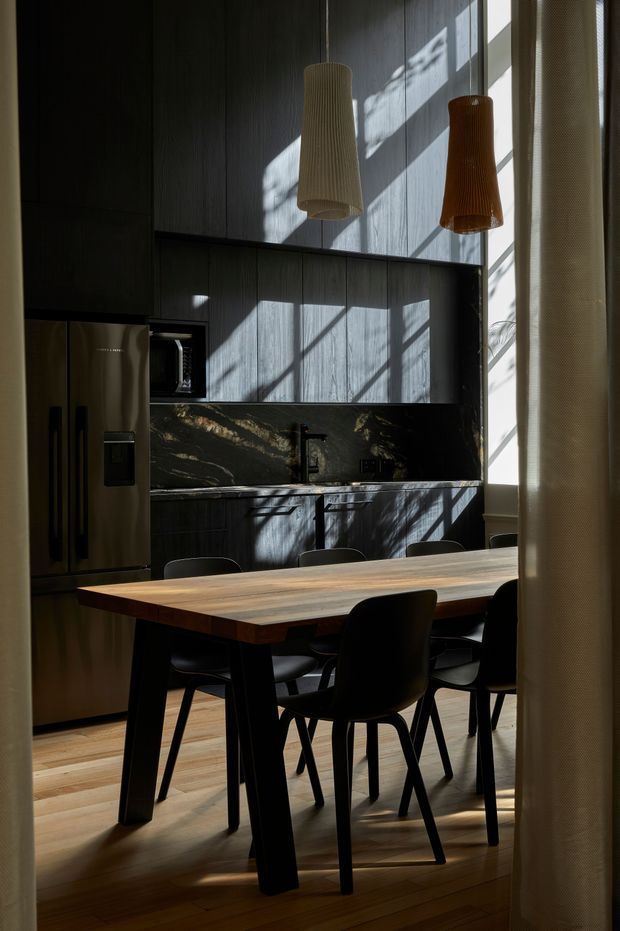 A moody, contemporary kitchen and dining area with dark vertically panelled cabinetry, a dramatic dark stone splashback, integrated stainless-steel refrigerator and microwave, and a timber dining table with black chairs. Two sculptural pendant lights hang above the table, while strong sunlight creates geometric shadows across the cabinetry and floor.