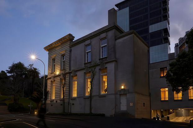 Dusk exterior view of a heritage-style masonry building with tall illuminated windows and classical facade detailing, integrated beside a much taller contemporary high-rise. The scene includes a landscaped urban streetscape, street lighting, and a recessed vehicle entrance at the adjoining modern structure.