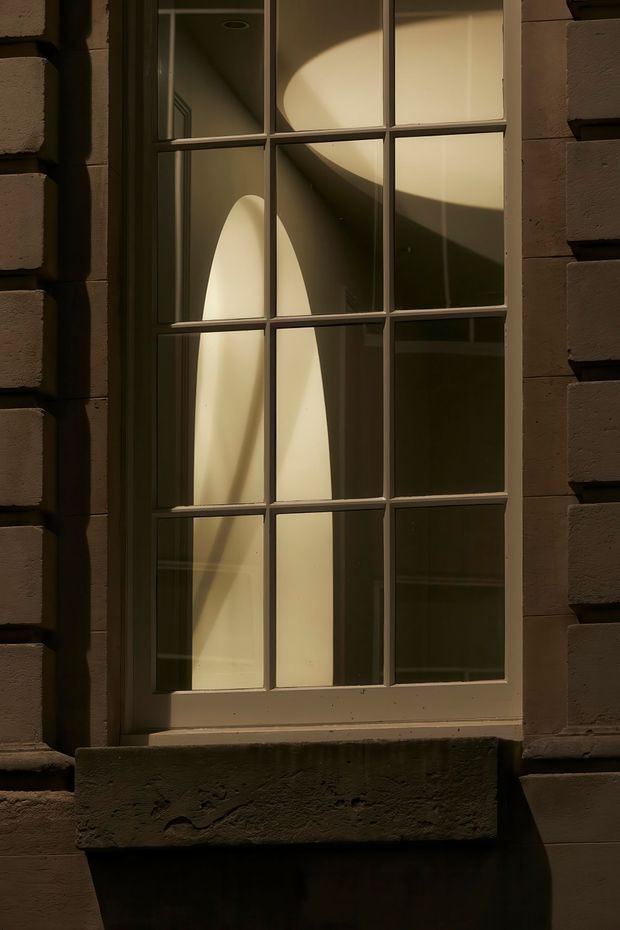Close-up exterior view of a tall, multi-pane window set into a dark masonry facade. Warm sunlight creates strong curved and diagonal patterns on the interior surfaces visible through the glazing, while the deep stone or rendered sill and surrounding blockwork emphasize the building envelope and traditional detailing.