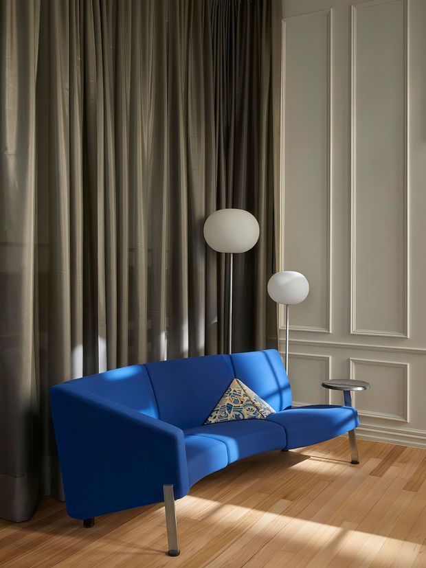 A refined living room with a vivid cobalt-blue modern sofa, a patterned accent cushion, two minimalist globe floor lamps, and a small side table. Floor-to-ceiling dark curtains frame a tall decorative wall with classic raised-panel moulding, while warm timber flooring and sunlight create a sophisticated, high-contrast atmosphere.